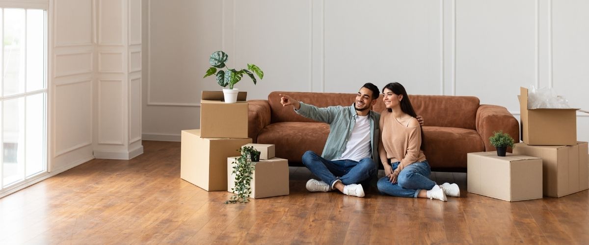 Un couple est assis sur un canapé, entouré de cartons, ce qui laisse supposer qu'ils sont en plein déménagement ou en train de déballer leurs affaires.
