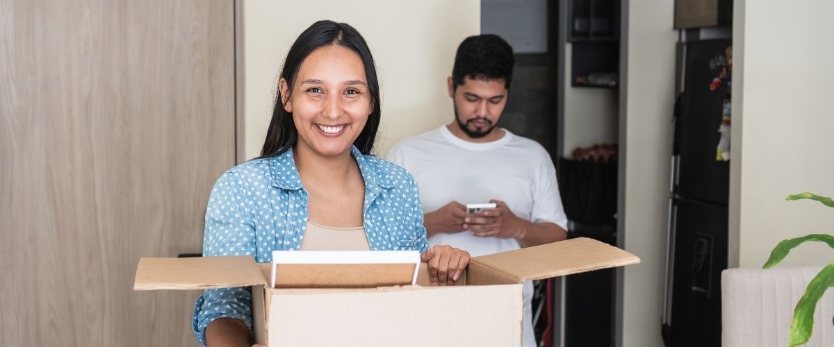 A woman holds a box while a man stands behind her, both smiling in a bright indoor setting.