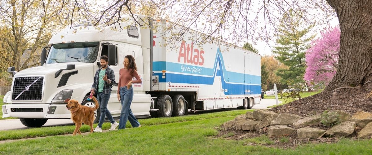 A couple strolls with their dog in front of a large truck, showcasing a casual outdoor scene.