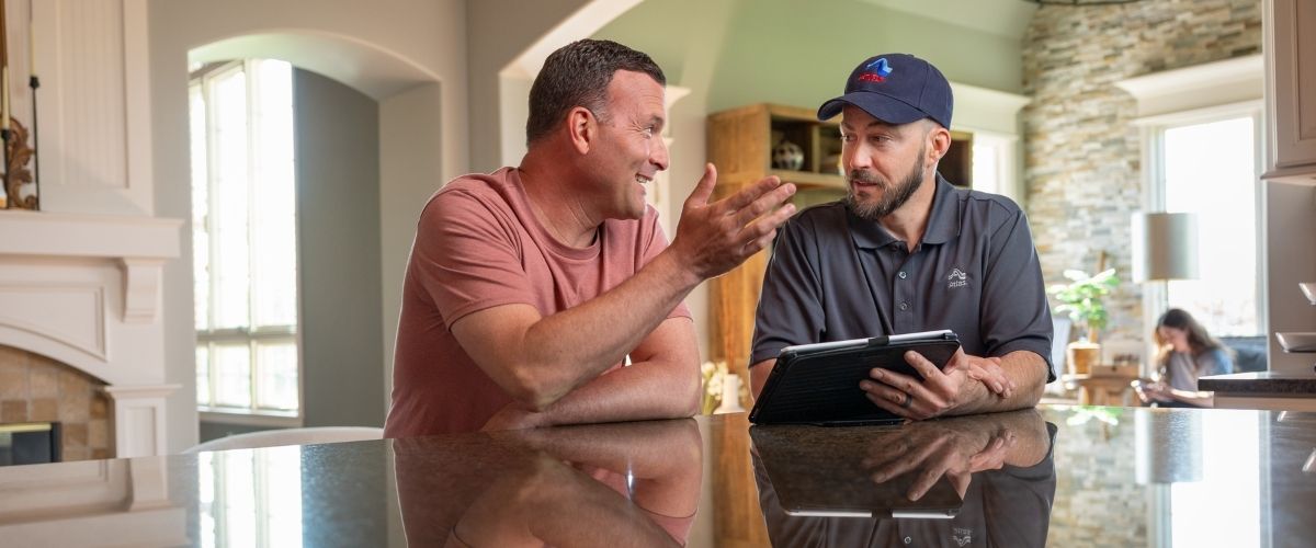 Two men seated at a kitchen table, engaged in discussion while looking at a tablet device.