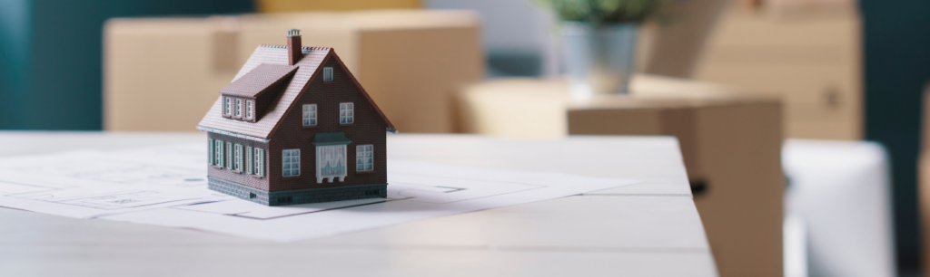 Une maison de poupée est posée sur une table, tandis qu'un homme déplace des cartons à l'arrière-plan.