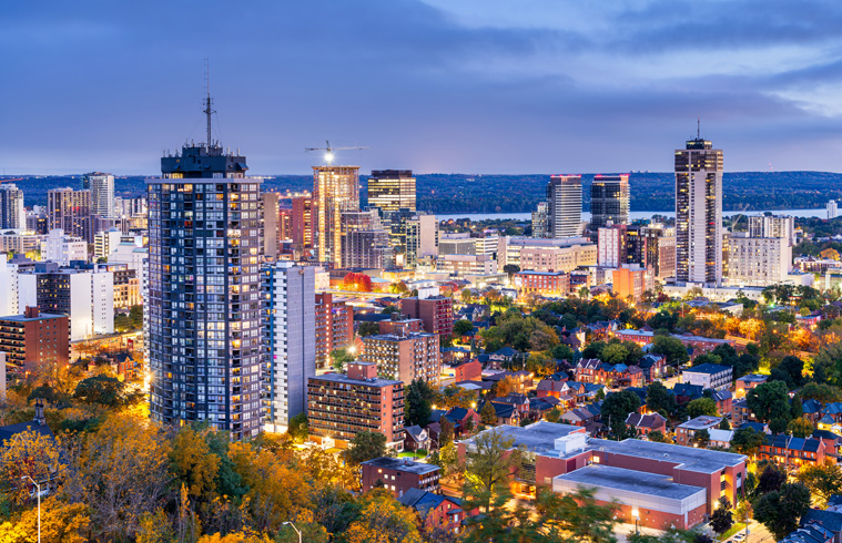 Evening view of Hamilton skyline with lit apartments, houses, autumn trees, and tall office towers at dusk.