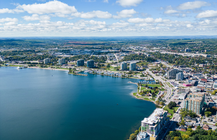 Aerial photo of Barrie, Ontario showing the lakeside waterfront, downtown high‑rise buildings, roads, and green spaces with hills.