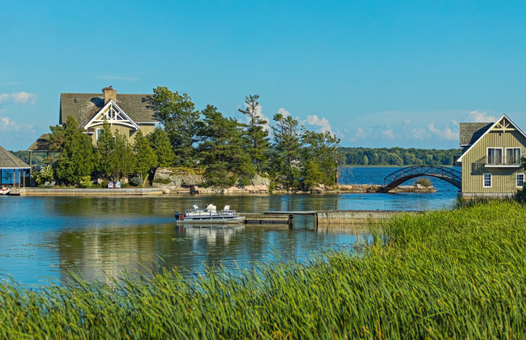 Maisons riveraines dans les Mille-Îles, près de Kingston, au Canada, avec pont en arc, bateau et roseaux sous un ciel dégagé.