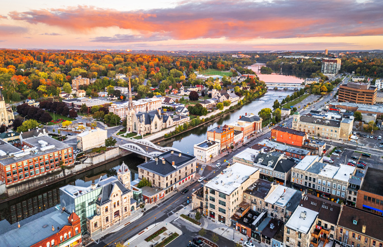 Sunset aerial view of Kitchener skyline with river, bridge, church steeple, and autumn trees.