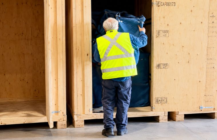 A man in a vest is lifting and moving a large cardboard box in a warehouse setting.