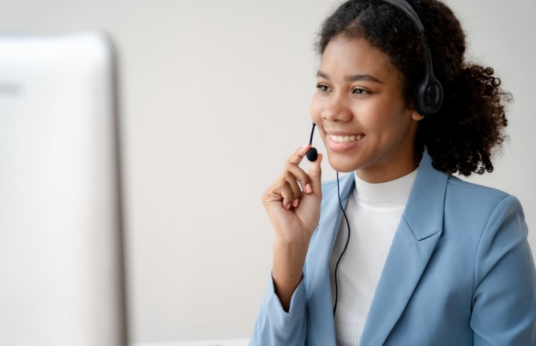 A woman with a headset smiles while working on a computer, indicating a positive and engaged work environment.
