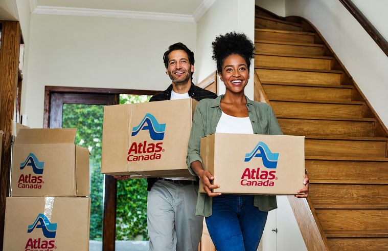 A man and woman are carrying boxes together inside a house, preparing for a move or organizing their belongings.
