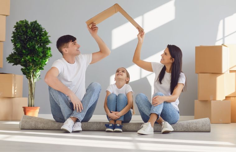 A family of four sits on the floor surrounded by cardboard boxes, smiling and enjoying their time together.