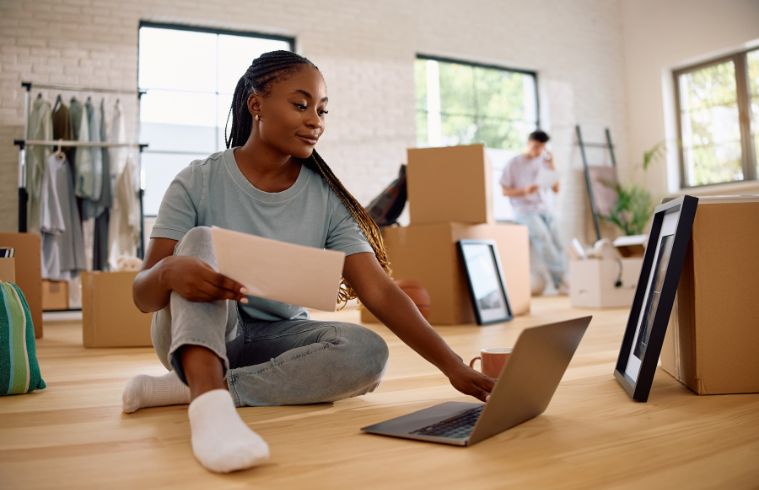 A woman sitting on the floor, using a laptop surrounded by cardboard boxes.