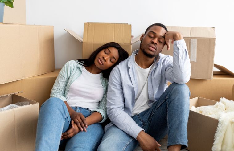 A young couple relaxes on the floor, surrounded by cardboard boxes, suggesting they are in the process of moving in.