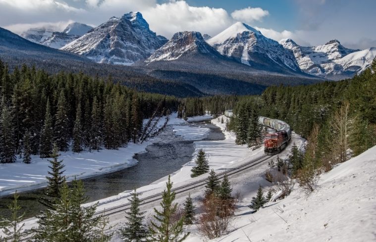 A train travels through snow-covered mountains, surrounded by tall, frosted trees under a clear blue sky.
